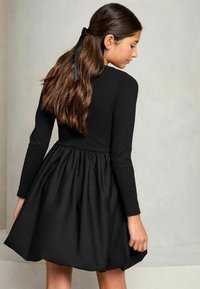 Schwarzes langärmliges Kleid mit einem gerippten Oberteil und gerafftem Rock. Die Haare sind mit einem schwarzen Band zurückgebunden. Glatter Stoff mit fließender Form.