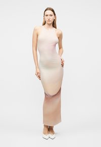 Nő, aki testhez simuló, ujjatlan, bézs-rózsaszín átmenetes hosszú ruhát és fehér hegyes cipőket visel, egyszerű világos háttér előtt áll.