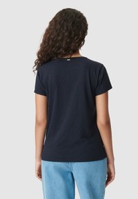 Mujer con cabello largo y rizado, vistiendo una camiseta básica de manga corta azul marino y jeans de mezclilla claros, vista desde atrás sobre un fondo gris.