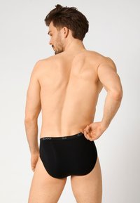 Schwarze Unterhose aus glattem Stoff. Mit einem elastischen Bund, auf dem der Markenname "SLOGGI" sichtbar ist, von hinten gezeigt.