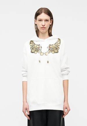 Frau trägt weißen Hoodie mit gestickten Tigerköpfen auf der Brust, kombiniert mit einem schwarzen Rock, steht vor einfachem Hintergrund.
