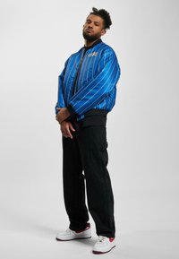 Blue satin bomber jacket with white pinstripes, ribbed cuffs, and FUBU logo, paired with black cargo pants and white sneakers with red accents.