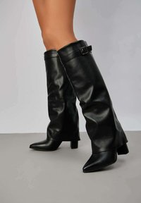 Bottes noires à talons hauts en cuir lisse, avec une tige large, un bout pointu et un accent en boucle sur le côté supérieur.