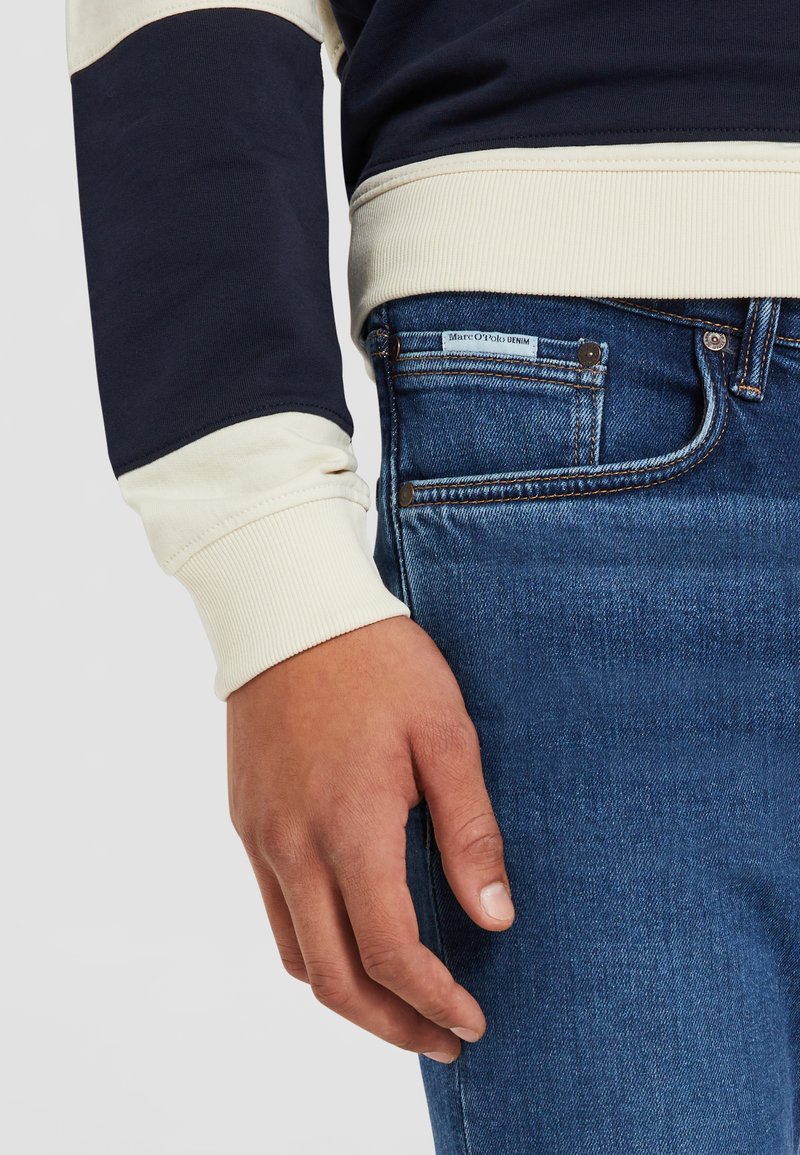 Sweat-shirt bleu marine et crème avec des poignets côtelés, associé à un jean en denim bleu foncé avec une étiquette sur la poche arrière. Main reposant sur le jean.