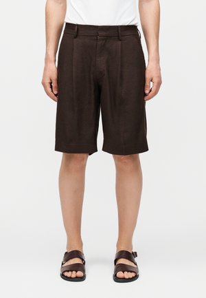 Άντρας που φοράει σκούρα καφέ, ραμμένα μέχρι το γόνατο σορτς και σκούρα καφέ δερμάτινα σανδάλια, στέκεται μπροστά σε ένα λευκό, μονόχρωμο φόντο.