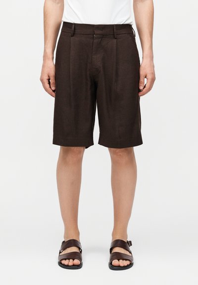 Homme portant un short marron foncé sur mesure jusqu'aux genoux et des sandales en cuir marron foncé, debout devant un fond blanc uni.