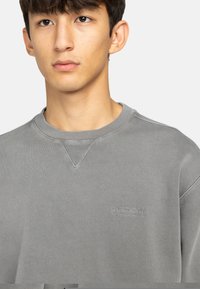 Grå sweatshirt med ribbet krave, en subtil V-udskæring og lille broderet logo på brystet. Blød tekstur og afslappet pasform.