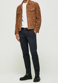 Hnědá semišová bunda se dvěma náprsními kapsami, bílý tričko, modré kalhoty a černé kotníkové boty. Hladká textury a přiléhavý střih.