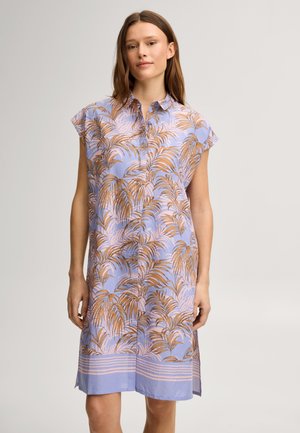 Femme portant une robe boutonnée mi-longue à manches courtes avec un fond bleu et un motif de feuilles de palmier marron.