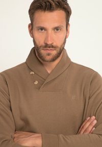 Maglione marrone con colletto a scialle, caratterizzato da due bottoni decorativi e un logo. Il tessuto sembra morbido con una superficie liscia.