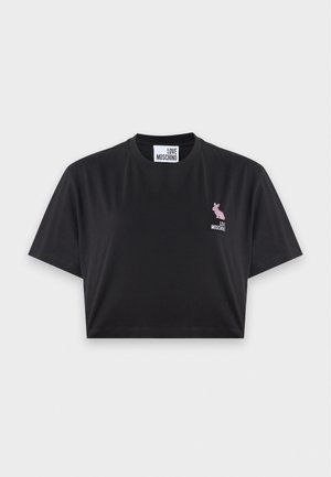 Femme debout, une main dans la poche, portant un t-shirt court noir, un jean bleu à jambes larges et des sandales noires à bout ouvert sur fond uni.