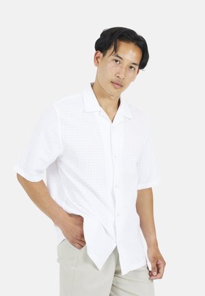 Młody mężczyzna o ciemnych włosach, ubrany w białą, krótkorękawową koszulę na guziki i jasnobeżowe spodnie, pozuje z jedną ręką w kieszeni.