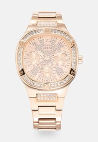 Montre Guess avec un bracelet en acier inoxydable rose doré, un cadran texturé avec des embellissements en cristal, et trois sous-cadrans pour la date et l'heure.