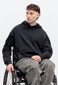 Jeune homme aux cheveux bouclés portant un sweat à capuche noir et un jean gris, assis en fauteuil roulant, regardant sur le côté sur un fond uni.