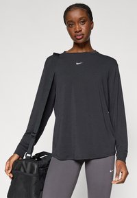 Μαύρο αθλητικό τοπ με μακριά μανίκια και στρογγυλή λαιμόκοψη, διακοσμημένο με μικρό λευκό λογότυπο Nike, συνδυασμένο με γκρίζες κολάν και μαύρη τσάντα. Απαλή ύφανση.