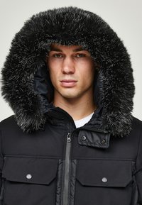 Svart polstret parkas med hette fôret med pels. Har glidelås, to brystlommer og en matt finish. Glinsende detaljer er synlige.