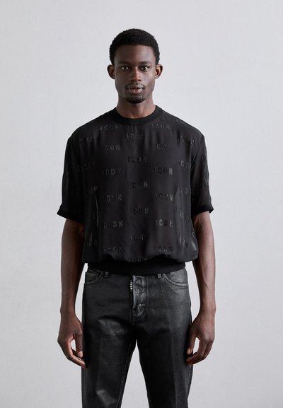 Chemise noire à manches courtes avec un motif texturé du mot "ICON" de différentes tailles, associée à un jean foncé ajusté.