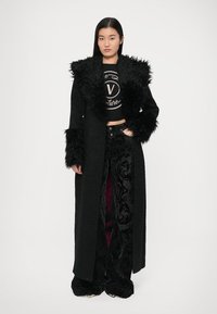 Črn dolg plašč z puhastim ovratnikom in manšetami, kombiniran s črno kratko majico in žametnimi hlačami z rožnim vzorcem.