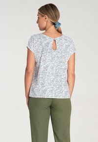 Biały top z krótkim rękawem z czarnym odręcznym wzorem, z wycięciem w kształcie klucza z tyłu. Zestawiony z zielonymi spodniami, podkreślającymi kształty obu elementów.