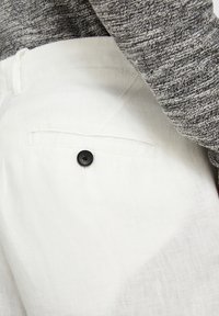 Vita linnesbyxor med en bakficka som har en enkel svart knapp. Tyget har en texturerad yta, i kombination med en grå, texturerad tröja.