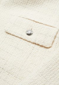 Crème textiel met een geweven patroon, met een zilveren knoop op een klepzak. Subtiele glinsteraccenten door het materiaal.