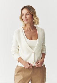 Cardigan crema con scollo a V, texture a coste e tre bottoni; abbinato a una canotta bianca e pantaloni beige. Palette di colori neutri.
