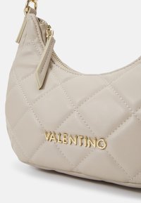 Vaaleanbeige verkkoleather-handrahka, jossa on kultasävyiset varusteet ja logo. Ominaisuuksina kaareva muoto ja vetoketjukiinnitys, jossa on pieniä kangaskoristeita.