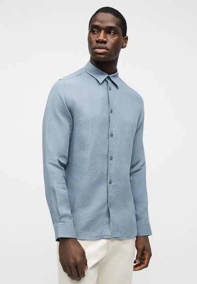 Jeune homme portant une chemise à manches longues bleu clair à boutons et un pantalon blanc, regardant vers sa droite sur un fond uni.