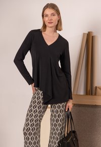 Schwarzes Langarmoberteil mit V-Ausschnitt, drapiertem Vorderteil und asymmetrischem Saum, kombiniert mit gemusterten, beige-schwarzen weiten Hosen.