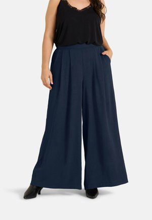 Vrouw die donkerblauwe wijde broek en een zwart mouwloos topje met kanten rand draagt, staand met één hand in de zak en zwarte hakken.