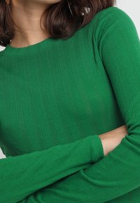 Grüner gerippter Pullover mit langen Ärmeln und Rundhalsausschnitt. Die Textur zeigt vertikale Streifen, die ein dezentes Muster bilden.