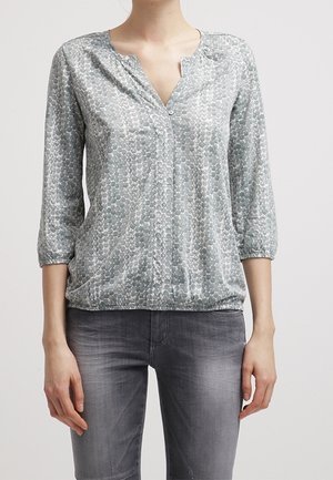 Femme portant un chemisier gris à motifs avec des manches trois-quarts et un jean gris délavé, debout devant un fond uni.