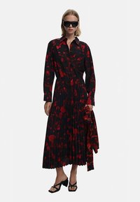 Juoda suknelė su raudonu gėlių raštu, ilgomis rankovėmis, klostuota sijonu ir sagotu apykakle. Dėvima su juodais akiniais nuo saulės ir batais su dirželiais.