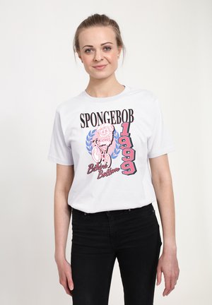 Giovane donna che indossa una maglietta bianca SpongeBob 1999 Bikini Bottom e pantaloni neri, in piedi contro uno sfondo bianco uniforme.