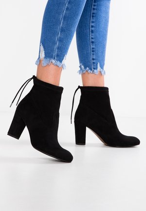 Sort ruskind støvler til anklerne med blokhæle, båret med blå skinny jeans med frynset kant på hvidt gulv.