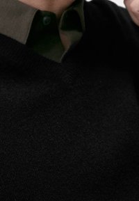 Czarny sweter dzianinowy z dekoltem w serek, noszony na ciemnozielonej koszuli z kołnierzykiem. Posiada gładką strukturę i ściągacz u dołu.