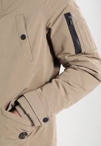 Giacca beige con finitura liscia. Presenta una tasca con zip sul maniche sinistro, due tasche sul petto con bottone e un polsino a costine.