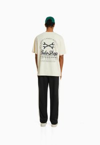 Short-sleeve cream t-shirt with a black graphic print and text on the back, paired with black track pants featuring white side stripes.