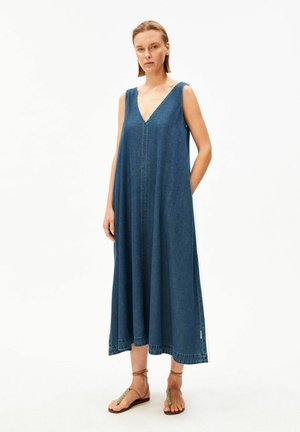 Femme portant une robe midi en jean ample sans manches à col en V avec des poches latérales et des sandales plates marron, debout devant un fond blanc.