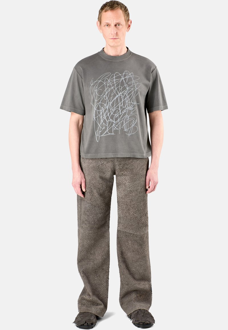 T-shirt gris avec un design abstrait en lignes blanches, associé à un pantalon ample gris texturé. Des chaussures plates noires complètent la tenue.