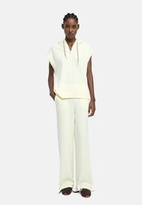 Kobieta stoi prosto w bezrękawniku w kolorze kremowym z zamkiem, dopasowanych szerokich spodniach oraz ciemnobrązowych sneakersach.