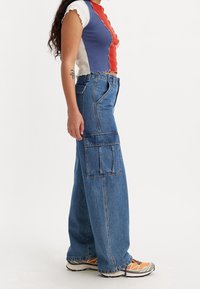 Person som bär högmidjade vida jeans cargobyxor, flerfärgad kort topp och orange sneakers, står i profil.