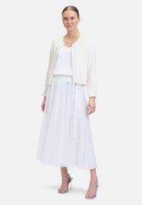 Weißer, texturierter Bomberjacke mit langen Ärmeln über einem weißen, ärmellosen Top. Fließender weißer Rock mit Taillenkrawatte und silbernen High-Heel-Sandalen.