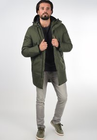 Olivegrønn parka med hette, med glidelås og trykknapper. Kombinert med grå jeans og grønne sneakers.