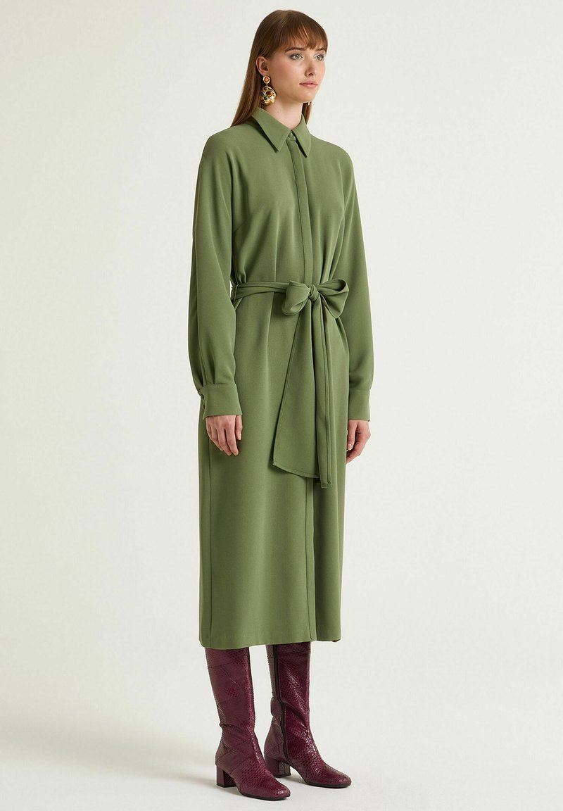 Grünes langärmliges Kleid mit Taillengürtel, kragenförmigem Ausschnitt, glatter Textur und geradem Schnitt. Kombiniert mit bordeauxroten kniehohen Stiefeln.