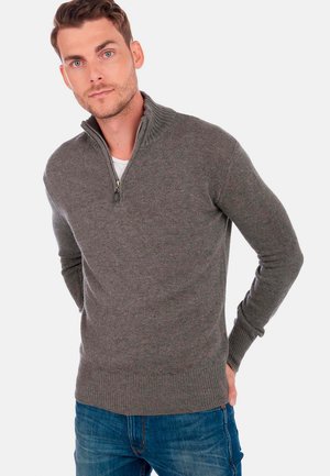 Pull en gris à zip avec col et poignets côtelés, fabriqué dans un matériau en maille texturée. Associé à un jean en denim bleu.