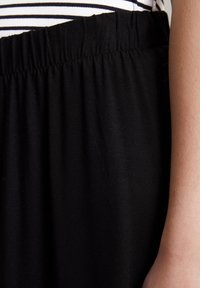 Sort, blødt stofskørt med elastisk talje. Glat tekstur, samlet ved taljen, vist med en stribet top i baggrunden.