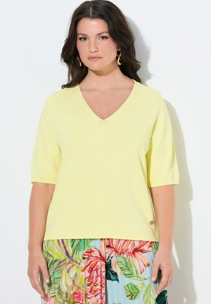 Frau mit dunkel gewelltem Haar, die goldene Creolen, ein hellgelbes V-Ausschnitt-Top und einen mehrfarbigen Blumenrock vor weißem Hintergrund trägt.