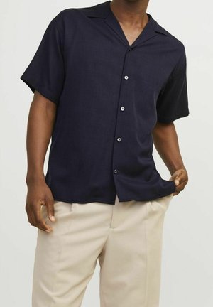 Mężczyzna w ciemnogranatowej koszuli na krótki rękaw z guzikami i jasnobeżowych spodniach, trzymający jedną ręką krawędź koszuli.