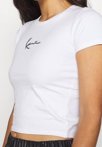 Modelka ve fitted bílém crop topu s černým podpisovým designem na hrudi, spárovaném s černými kalhotami s elastickým pasem.